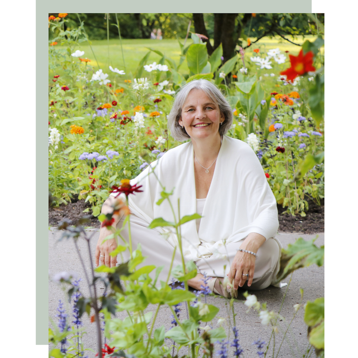 Energetikerin Christine Gningue lächelt im Schneidersitz inmitten eines bunten Blumenbeets.