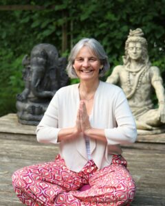 Energetikerin Christine Gningue sitzt im Meditationssitz mit zusammengelegten Händen vor Ganesha- und Shiva-Statuen.